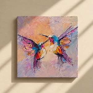 May include: Square canvas art featuring two hummingbirds in a textured oil painting style. The birds are depicted with vibrant colors, including purple, blue, orange, and white, against a soft pink and peach background.