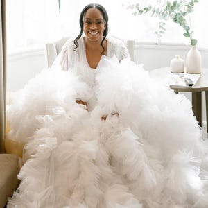 May include: A smiling person wearing a white wedding dress with a voluminous, ruffled skirt. The dress has a V-neckline and is adorned with delicate embellishments. The image is taken indoors, with natural light illuminating the scene.