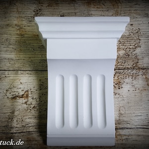 Könnte beinhalten: Ein weißes architektonisches Kragstein mit klassischem Design. Das Kragstein hat eine rechteckige Form mit detaillierter Oberseite und einer vertikalen gerillten Platte. Der Hintergrund ist eine rustikale Holzoberfläche. Die Website-Adresse "luxstuck.de" ist unten links sichtbar.