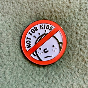 May include: A round enamel pin with a red border and a red diagonal line through a white circle. The white circle features a cartoon face and the words "NOT FOR KIDS" in black.