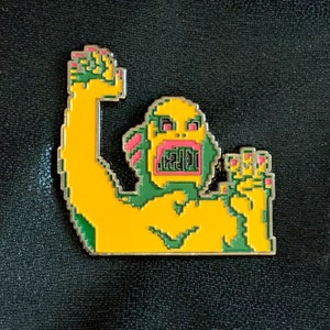 Könnte beinhalten: Ein gelber Emaille-Pin mit einem pixeligen Monster mit erhobenem Arm und offenem Mund. Das Monster hat grüne und rosa Akzente und einen silbernen Umriss. Der Pin ist vor einem schwarzen Hintergrund platziert.