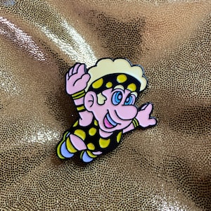 Pode incluir: Um broche de esmalte com um personagem de desenho animado com uma faixa de cabeça amarela, olhos pretos e uma roupa com manchas pretas e amarelas.