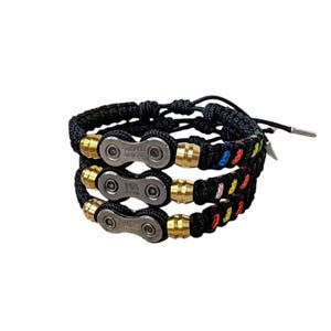 May include: Three black braided bracelets with metal accents. Each bracelet features a bicycle chain link design with gold-coloured metal beads and chain links. One bracelet has colourful beads. The bracelets are adjustable.