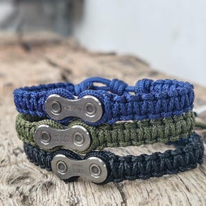 Könnte beinhalten: Drei vertikal gestapelte Armbänder, die jeweils ein silberfarbenes Fahrradkettenglied als Mittelstück in einem geflochtenen Kordelband aufweisen. Das obere Band ist blau, das mittlere olivgrün und das untere Band ist schwarz.