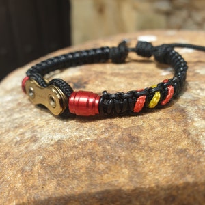 Könnte beinhalten: Ein schwarzes Paracord-Armband mit einem goldenen Kettenglied und einem roten und gelben Akzent. Das Armband liegt auf einer braunen Oberfläche.