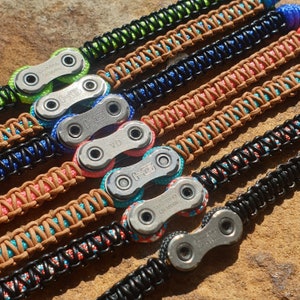 Op de afbeelding: Een set van zes armbanden met verschillende kleuren paracord en een zilveren fietskettinglink. De armbanden zijn geweven met verschillende kleuren, waaronder zwart, groen, blauw, rood en bruin.
