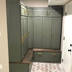 May include: A corner entryway with a built-in bench and storage cabinets. The cabinets are painted a dark green color and have gold hardware. The bench is made of wood and has a dark brown finish. The floor is made of brick tiles in a herringbone pattern.