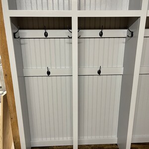 K&ouml;nnte beinhalten: Ein wei&szlig;er Holz-Schrank mit zwei Abteilen, jedes mit einem Regal und zwei Reihen Haken. Der Schrank ist wei&szlig; lackiert und hat eine Holzmaserung. Die Haken sind aus schwarzem Metall.