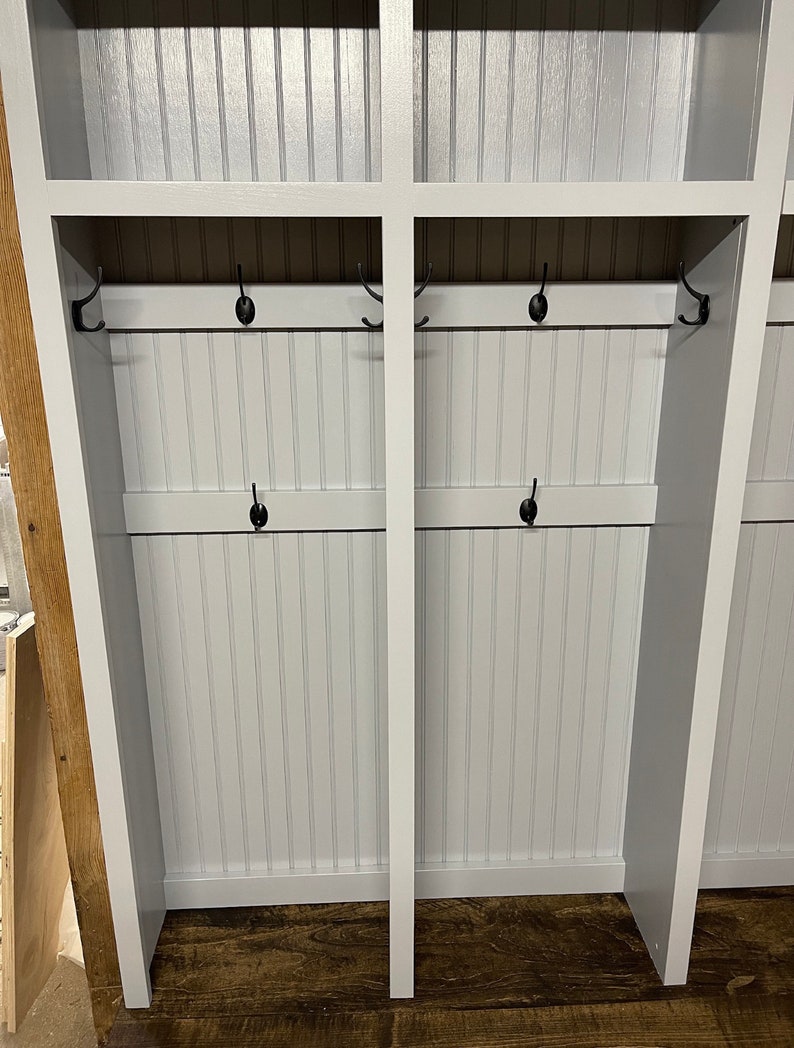 K&ouml;nnte beinhalten: Ein wei&szlig;er Holz-Schrank mit zwei Abteilen, jedes mit einem Regal und zwei Reihen Haken. Der Schrank ist wei&szlig; lackiert und hat eine Holzmaserung. Die Haken sind aus schwarzem Metall.