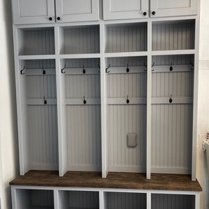 K&ouml;nnte beinhalten: Eine graue Holzbank f&uuml;r den Eingangsbereich mit drei F&auml;chern und einem oberen Regal mit drei T&uuml;ren. Die Bank hat eine braune Holzplatte und schwarze Haken an den Seiten.