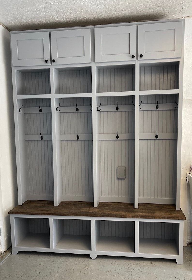 K&ouml;nnte beinhalten: Eine graue Holzbank f&uuml;r den Eingangsbereich mit drei F&auml;chern und einem oberen Regal mit drei T&uuml;ren. Die Bank hat eine braune Holzplatte und schwarze Haken an den Seiten.
