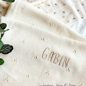 May include: A white knitted baby blanket with a beige embroidered name "GABIN" and the text "La broderie - Police #5 Beige".