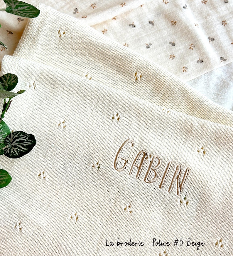 May include: A white knitted baby blanket with a beige embroidered name "GABIN" and the text "La broderie - Police #5 Beige".