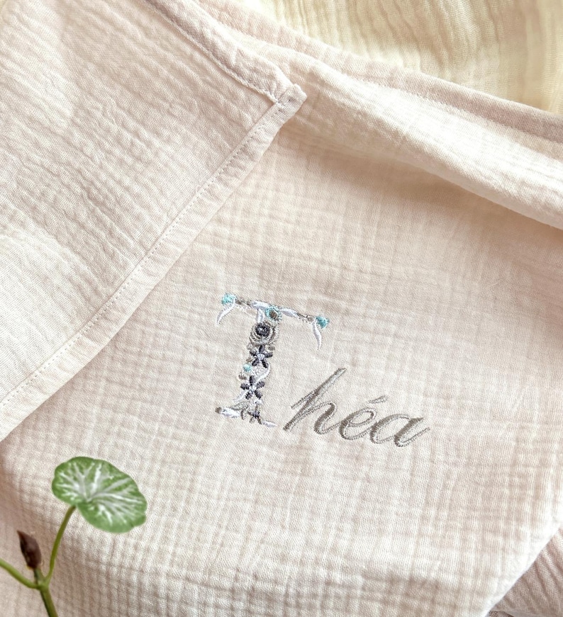 May include: A white muslin baby blanket with the name "Th&eacute;a" embroidered in gray thread. The letter "T" is decorated with small floral details.