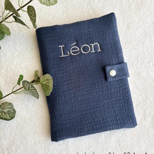 May include: A navy blue fabric cover with the name "L&eacute;on" embroidered in white. The cover has a button closure and the text "La broderie: Police #2 Argent" is at the bottom.
