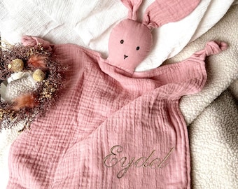 May include: A pink baby security blanket with a bunny head and long ears. The blanket has a textured, crinkled appearance and the name "Eydel" is embroidered in gold thread. The bunny's face has black embroidered eyes and a small nose.