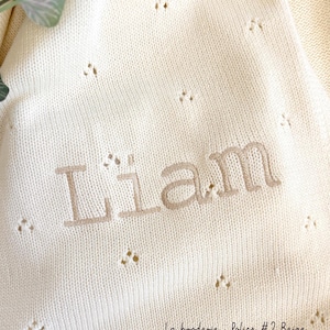 May include: A white knitted baby blanket with a beige embroidered name "Liam" and the text "La broderie Police #2 Beige".