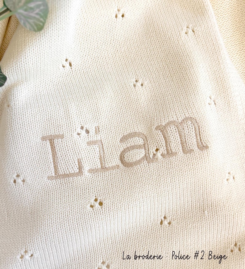 May include: A white knitted baby blanket with a beige embroidered name "Liam" and the text "La broderie Police #2 Beige".