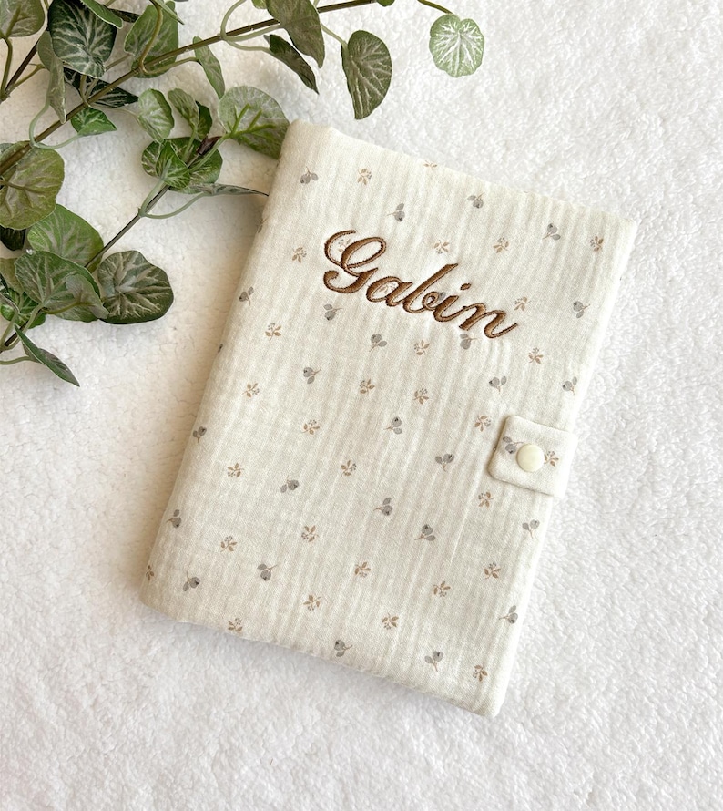 May include: A beige muslin baby book with a white button closure and a brown embroidered name "Galbin" on the front.
