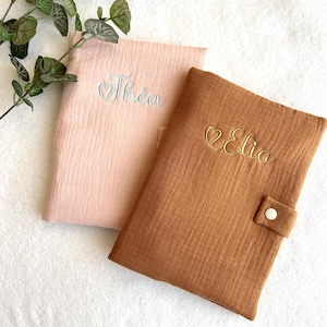 May include: Two personalized baby book covers. One is light pink with "Th&eacute;a" embroidered in light blue, and the other is brown with "Elio" embroidered in gold. Both covers have a textured, crinkled appearance.