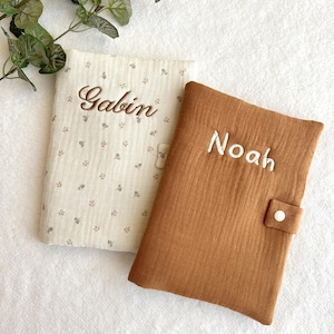 May include: Two personalized baby book covers. One is beige with a floral pattern and the name "Gabin" embroidered in brown. The other is brown with the name "Noah" embroidered in white. Both have a button closure.