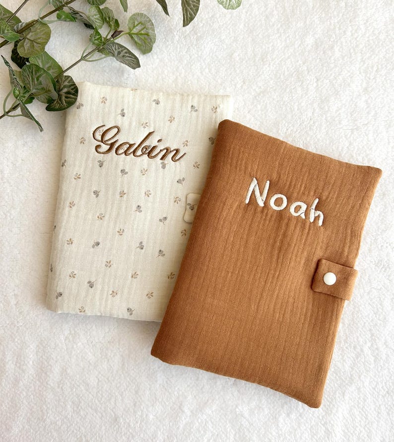 May include: Two personalized baby book covers. One is beige with a floral pattern and the name "Gabin" embroidered in brown. The other is brown with the name "Noah" embroidered in white. Both have a button closure.