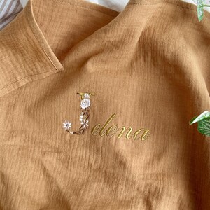 May include: A brown muslin baby blanket with the letter "J" embroidered in gold thread with white flowers and the name "Elena" embroidered in gold thread.