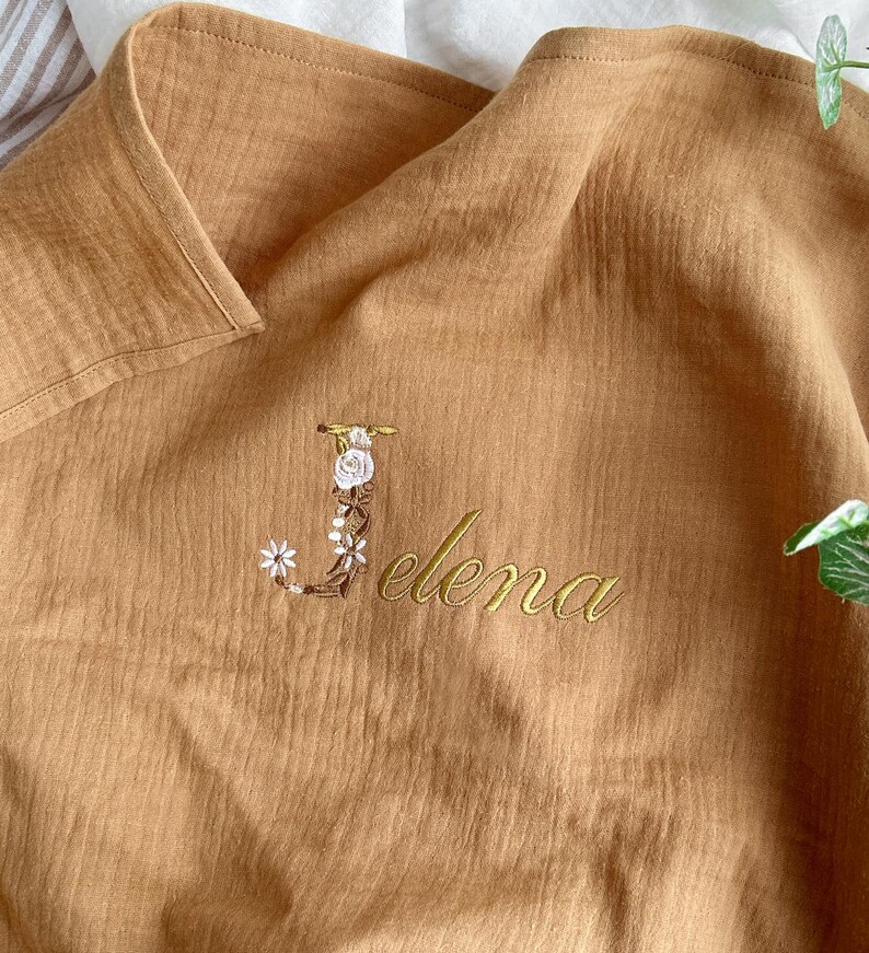 May include: A brown muslin baby blanket with the letter "J" embroidered in gold thread with white flowers and the name "Elena" embroidered in gold thread.