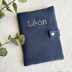 May include: A dark blue fabric baby book cover with a white button closure. The cover is personalized with the name "L&eacute;on" embroidered in white thread. The text "La broderie : Police #2 Argent" is printed in black at the bottom of the image.