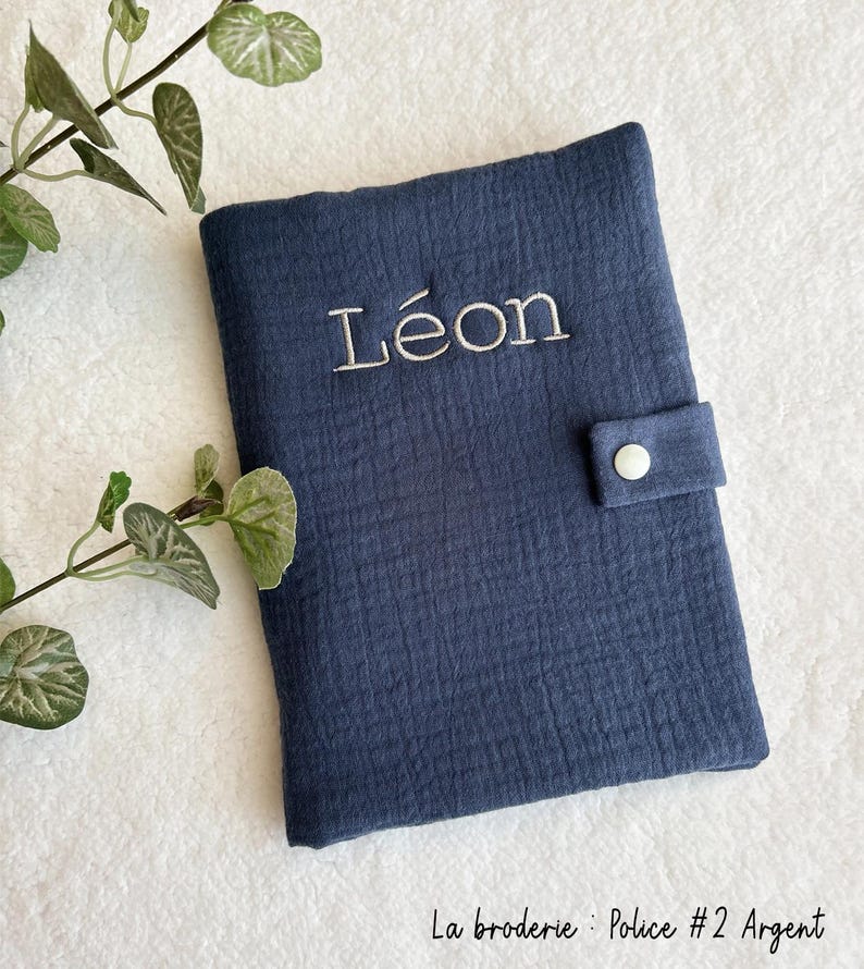 May include: A dark blue fabric baby book cover with a white button closure. The cover is personalized with the name "L&eacute;on" embroidered in white thread. The text "La broderie : Police #2 Argent" is printed in black at the bottom of the image.