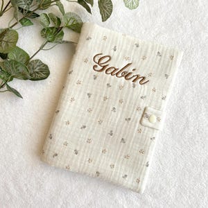 May include: Cream-colored baby book cover with the name "Gabin" embroidered in brown thread. The cover features a delicate floral pattern in shades of gray and brown, and a button closure.