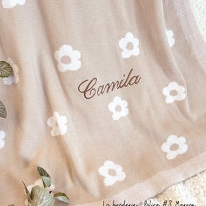 May include: A beige knitted baby blanket with a white daisy pattern and the name "Camila" embroidered in brown thread. The blanket has a brown border and the text "La broderie: Police #3 Marron" is printed in the bottom right corner.