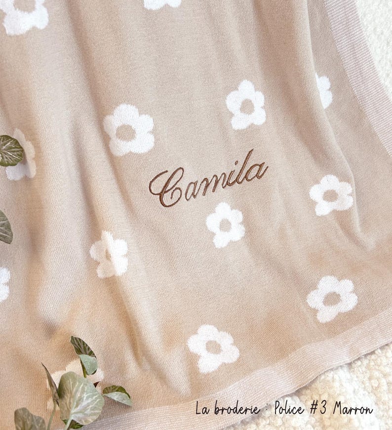 May include: A beige knitted baby blanket with a white daisy pattern and the name "Camila" embroidered in brown thread. The blanket has a brown border and the text "La broderie: Police #3 Marron" is printed in the bottom right corner.