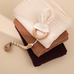 May include: A stack of three muslin swaddles in beige, brown, and burgundy. A wooden teether with a white bunny ear design is on top of the swaddles. A wooden bead chain with a clip is in the foreground.