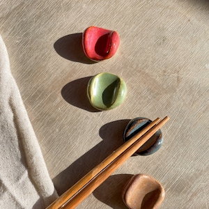 May include: Five ceramic chopstick rests in different colours and patterns. The rests are arranged in a vertical line on a light brown surface. A pair of wooden chopsticks are lying on the surface.