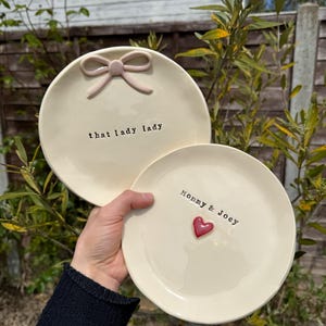 May include: Two cream ceramic plates. One features a pink bow and the text "that lady lady". The other has a red heart and the text "Mommy & Joey".