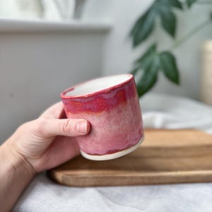 May include: A handmade ceramic mug with a vibrant red and pink ombre glaze. The mug has a white interior and a slightly textured surface. The mug is held in a hand, showcasing its size and shape.