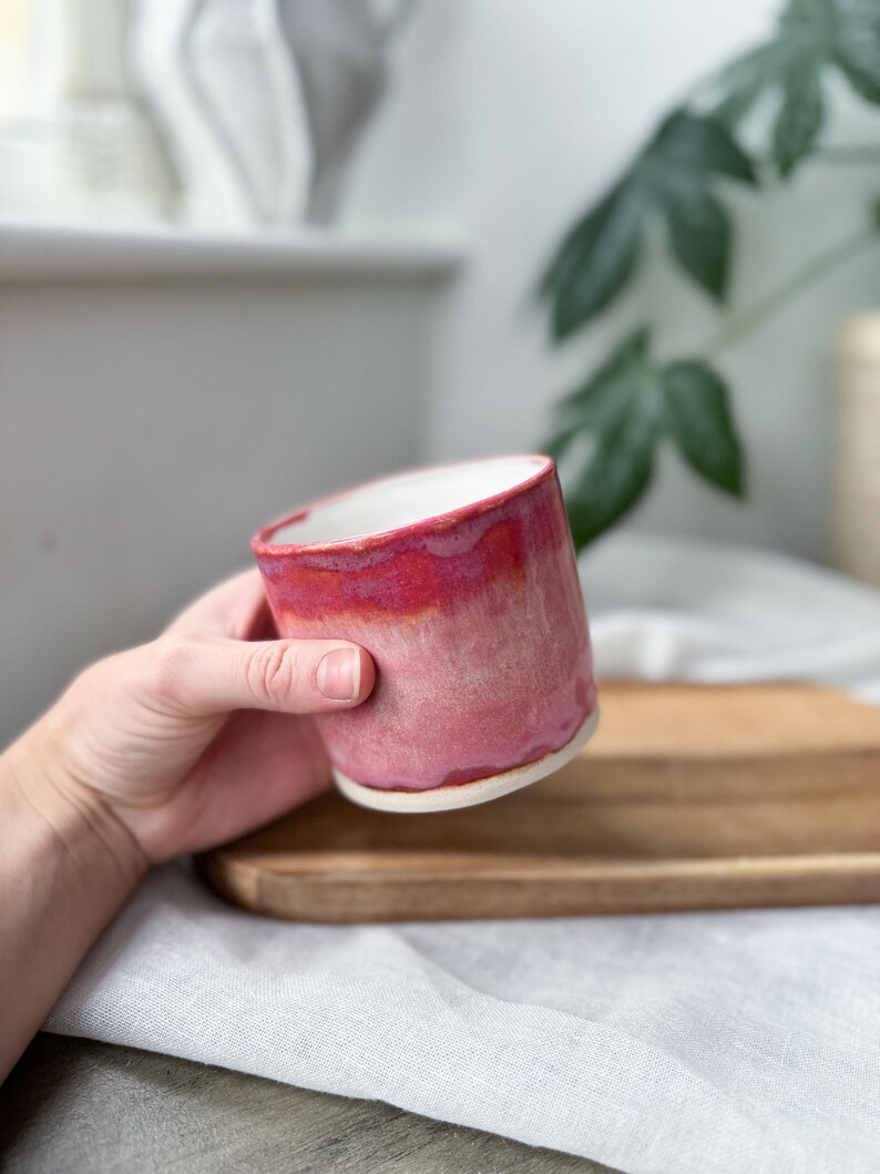 May include: A handmade ceramic mug with a vibrant red and pink ombre glaze. The mug has a white interior and a slightly textured surface. The mug is held in a hand, showcasing its size and shape.