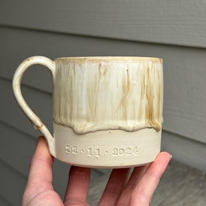 以下が含まれることがあります： クリーム色のセラミックマグカップ。茶色の釉薬が垂れたようなデザイン。丸みを帯びた取っ手があり、底には日付「22.11.2024」が刻印されています。手で持たれています。
