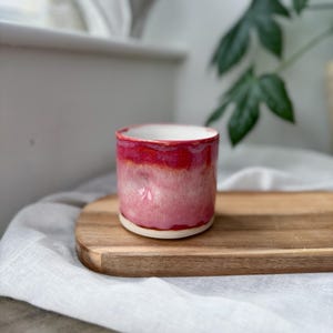 May include: A handmade ceramic cup with a vibrant red and pink glaze. The cup has a cylindrical shape with a white interior and sits on a wooden tray. The cup is a unique piece of pottery, perfect for coffee or tea.