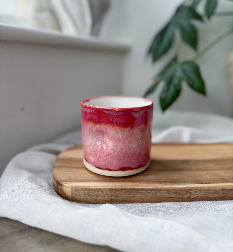 May include: A handmade ceramic cup with a vibrant red and pink glaze. The cup has a cylindrical shape with a white interior and sits on a wooden tray. The cup is a unique piece of pottery, perfect for coffee or tea.