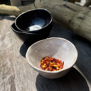 May include: Three handmade ceramic bowls. Two are stacked, with a glossy black finish. The third bowl is off-white and contains red pepper flakes. The bowls are on a weathered wooden surface.