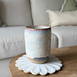 Könnte beinhalten: Ein zylindrischer Keramikbecher mit einer gesprenkelten, cremeweißen, blauen und braunen Glasur, der auf einem weißen, blumenförmigen Keramikuntersetzer steht. Der Becher hat einen glatten, abgerundeten Rand und einen natürlichen, unglasierten Boden.