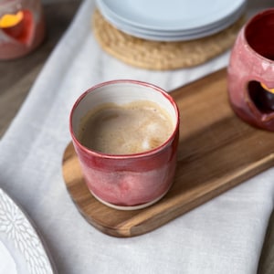 Op de afbeelding: Een rode keramische mok gevuld met koffie staat op een houten serveerplank. De mok heeft een witte binnenkant en een rode buitenkant. Een stapel witte borden en een geweven onderzetter staan op de achtergrond.