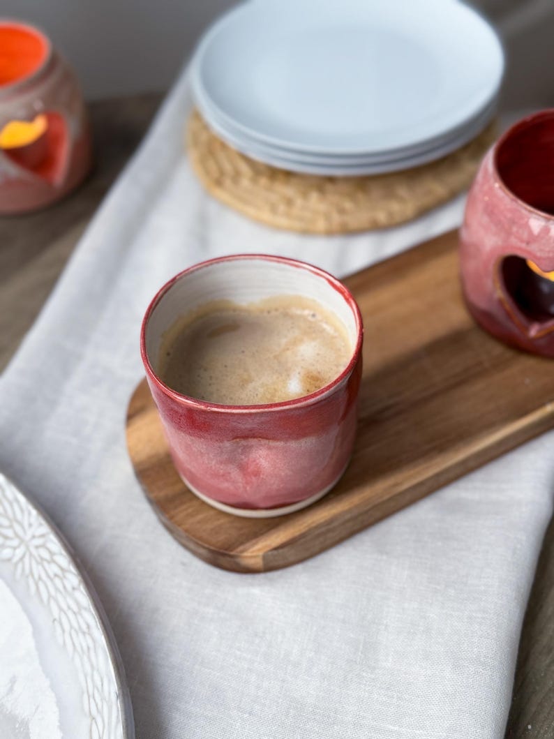 May include: A red ceramic mug filled with coffee sits on a wooden serving board. The mug has a white interior and a red exterior. A stack of white plates and a woven coaster are in the background. A white cloth is underneath the mug and board.