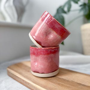 May include: Two handmade ceramic cups with a glossy, pink and red glaze. The cups are stacked on top of each other, showcasing their unique shape and design. The cups have a white base and a smooth, polished finish.