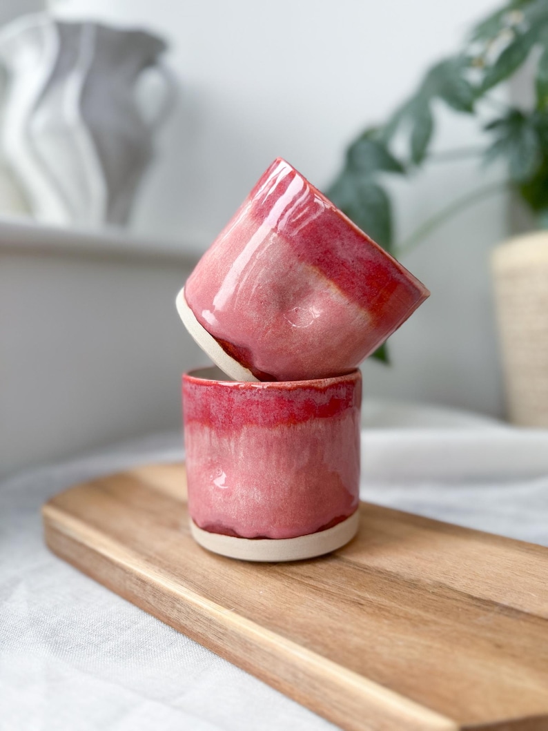 May include: Two handmade ceramic cups with a glossy, pink and red glaze. The cups are stacked on top of each other, showcasing their unique shape and design. The cups have a white base and a smooth, polished finish.