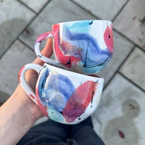 Peut inclure: Deux tasses en céramique blanche avec des motifs aquarelles abstraits en rouge, bleu et turquoise. Chaque tasse a une anse incurvée et un motif peint unique. Les tasses sont tenues dans la main d'une personne.