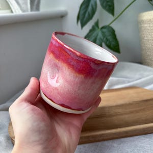 May include: A handmade ceramic mug with a gradient of pink and red hues. The interior is white, and the mug has a slightly angled rim. The mug is held in a hand, showcasing its size and shape. The mug is likely used for drinking beverages.