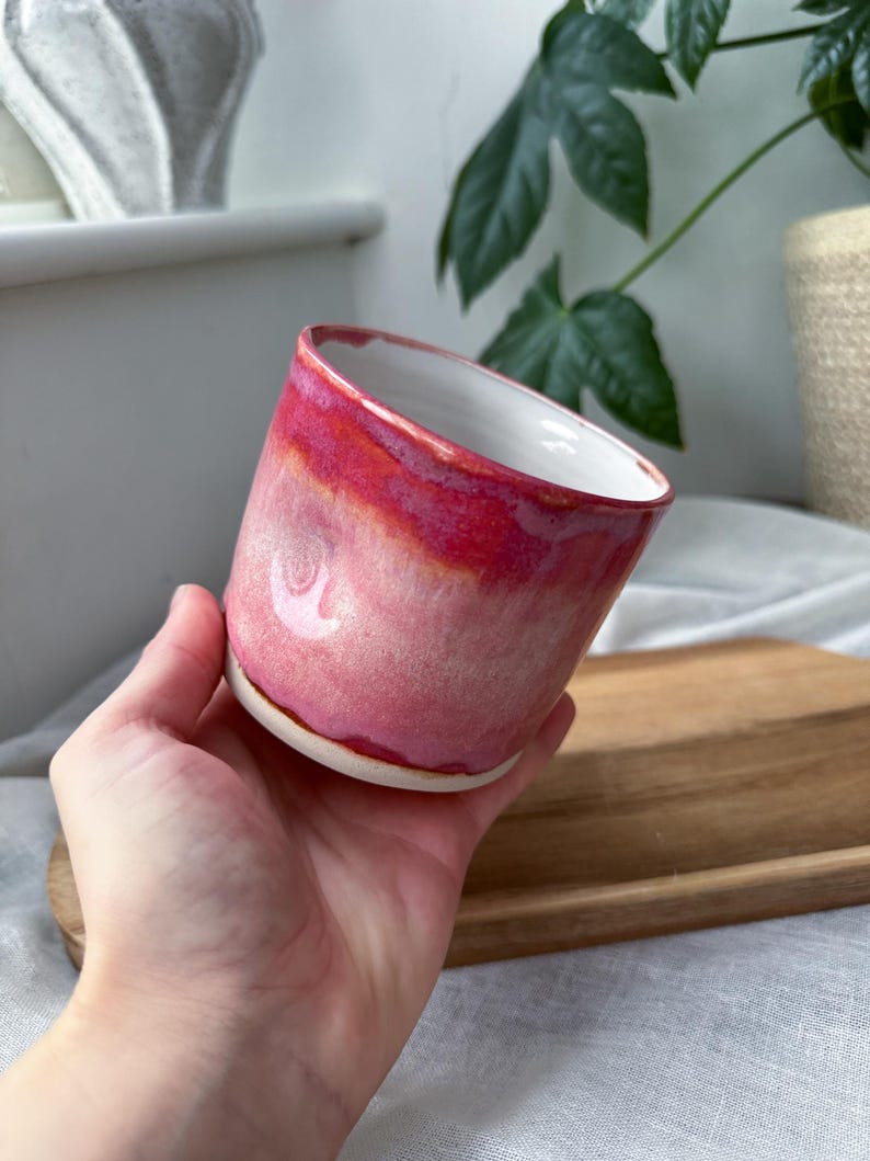 May include: A handmade ceramic mug with a gradient of pink and red hues. The interior is white, and the mug has a slightly angled rim. The mug is held in a hand, showcasing its size and shape. The mug is likely used for drinking beverages.